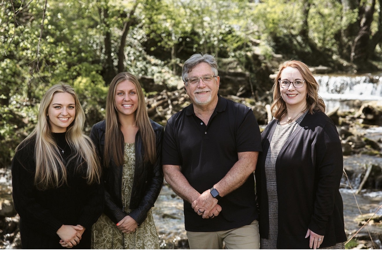 Four adults standing outdoors by a forest stream with a small waterfall, smiling at the camera.