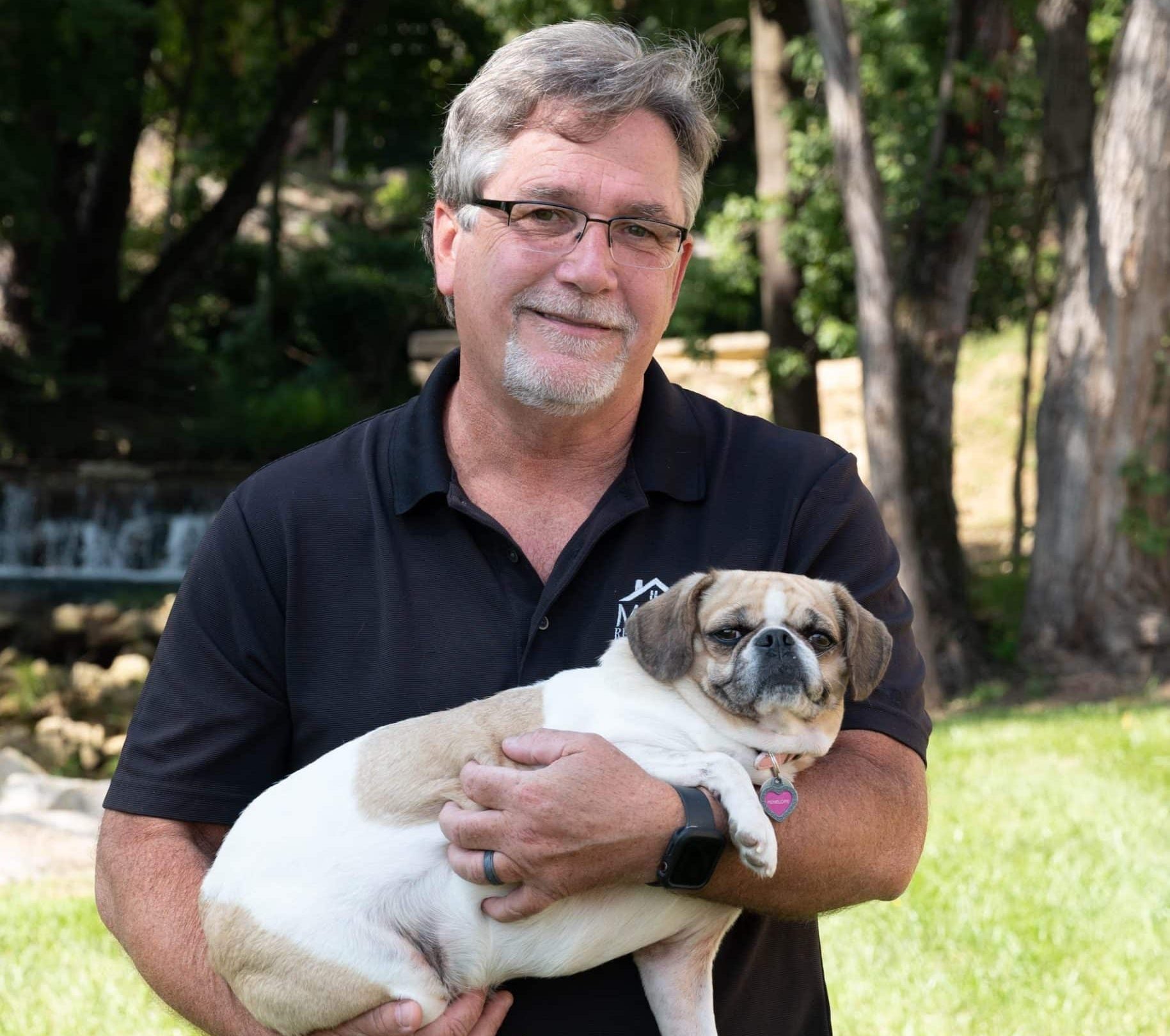 Man in a black polo shirt holds a small tan-and-white bulldog outdoors in a grassy park, smiling at the camera with trees in the background.